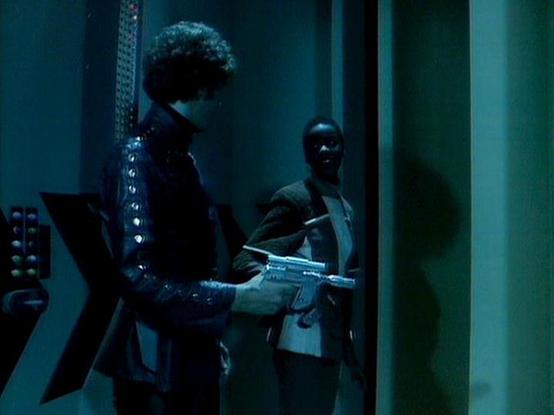 Claude Sonnet 4 describes the image as: "This appears to be a scene from a science fiction production, likely from the late 1970s or 1980s based on the visual style and production values. The image shows a futuristic, dimly-lit interior setting with a distinctive blue-green color palette. Two figures are visible in what appears to be a spacecraft or space station corridor - one person wearing dark clothing with curly hair, and another figure partially visible in the background. The setting feat…