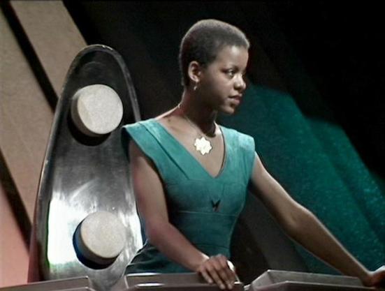 Claude Sonnet 4.5 describes the image as: "This image captures a scene from what appears to be a science fiction television production, likely from the 1980s based on the visual style and set design. The setting features a futuristic control panel or console with sleek, curved metallic elements in the background, characteristic of sci-fi aesthetics of that era.
The actress Josette Simon is shown in profile, wearing a striking teal-colored sleeveless dress with a V-neck design and a decorative …