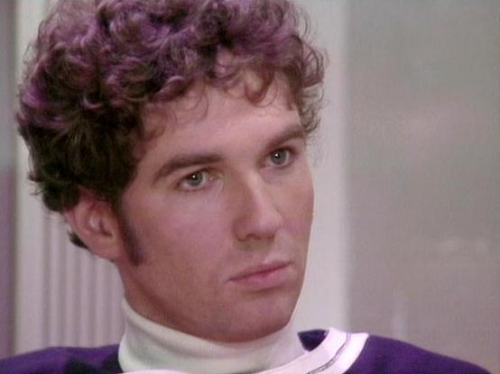 Claude Haiku 4.5 describes the image as: "# Image Description

This image shows a person with distinctive curly purple-tinted hair styled in a voluminous, permed fashion characteristic of 1980s television aesthetics. The individual is wearing a purple and white collared garment against a plain, neutral background with soft lighting typical of indoor studio settings.

The photograph appears to be a professional headshot or promotional image from a television production, given its quality and fra…