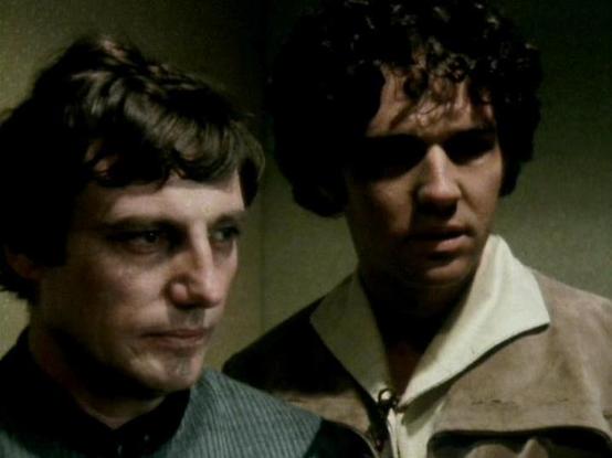 Claude Sonnet 4.5 describes the image as: "This appears to be a scene from the British science fiction television series "Blake's 7," which aired from 1978 to 1981. The image shows two characters in what appears to be a dimly lit, confined interior space, likely aboard a spacecraft or station. The lighting creates a tense, dramatic atmosphere with shadows falling across their faces.

The character on the left wears dark clothing with what appears to be a ribbed or textured collar detail. The ch…