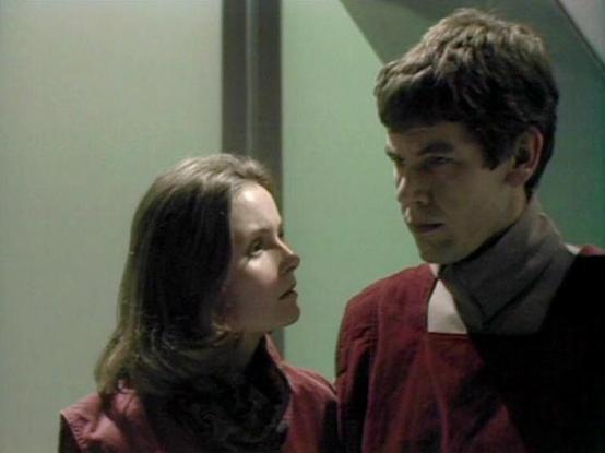 Claude Haiku 4.5 describes the image as: "# Scene Description

This image shows two actors in what appears to be a sci-fi setting, likely from a television production. They are positioned in an indoor corridor or hallway with pale green-tinted walls and neutral lighting typical of a spaceship or futuristic facility interior.

The actor on the right wears a distinctive red and black uniform with a high collar, characteristic of Starfleet-style costumes. He has dark hair and is looking toward his…