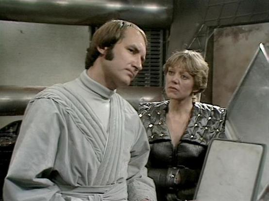 Claude Sonnet 4.5 describes the image as: "This image appears to be from a science fiction television production, likely from the late 1970s or early 1980s based on the visual style and production design. The setting is a futuristic spacecraft or station interior, characterized by industrial metallic pipes, utilitarian wall panels, and a functional, somewhat austere aesthetic typical of the era's sci-fi productions.

The scene shows two characters in a serious moment of dialogue or contemplatio…