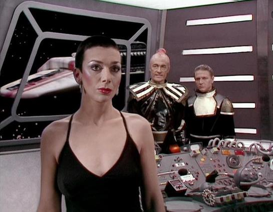 Claude Sonnet 4 describes the image as: "This appears to be a scene from a science fiction television production, likely from the 1970s or 1980s based on the aesthetic and production values. The setting is a futuristic spacecraft interior with characteristic sci-fi design elements including control panels with various buttons and displays, metallic surfaces, and a window showing stars and what appears to be another spacecraft in the background.

The scene shows three people in what appears to b…