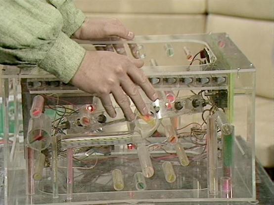 Claude Sonnet 3.7 describes the image as: "The image shows hands interacting with a transparent acrylic or plastic device that appears to be some kind of experimental apparatus or musical instrument. The hands, wearing a light green or mint-colored sleeve, are manipulating controls or components inside the clear box, which contains various tubes, wires, and electronic elements. Some small red lights are visible inside the device. The apparatus has multiple cylindrical components arranged inside…