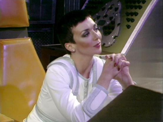 Claude Sonnet 3.7 describes the image as: "The image shows a person with short dark hair and makeup in what appears to be a futuristic or sci-fi setting. They're wearing a white outfit with light-colored details and are seated with their hands clasped together, looking thoughtfully to the side. The background features yellow/gold panels and what looks like some technical equipment or control panels with buttons, suggesting this is from a science fiction production. The aesthetic has a distincti…