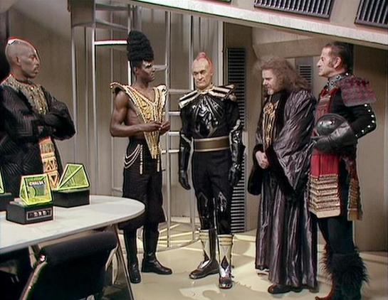 Claude Haiku 4.5 describes the image as: "# Scene Description

This image appears to be from a science fiction television production, likely from the 1970s or 1980s based on the styling and video quality. The setting is a futuristic interior space with metallic paneling and institutional-style architecture typical of space stations or starship corridors.

The scene depicts six individuals in elaborate costumes gathered in what appears to be an official or ceremonial meeting. The costumes range …