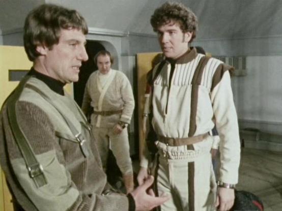 Claude Sonnet 4.5 describes the image as: "This image appears to be from a science fiction television production, showing three men in a futuristic corridor setting. The scene features utilitarian, industrial design elements typical of 1970s-80s sci-fi aesthetics, with grey walls and functional architecture.

In the foreground, two men are engaged in conversation. The person on the left wears a tactical-style vest in beige/tan tones with visible straps and pouches, paired with a dark turtleneck…