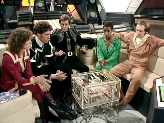 Claude Sonnet 4 describes the image as: "This appears to be a scene from the British science fiction television series "Blake's 7," set aboard the spacecraft Liberator. Five crew members are gathered in the ship's main lounge area, seated on the characteristic beige/cream colored seating. They're positioned around what appears to be a transparent container or device on the central table. The setting features the show's distinctive futuristic interior design with metallic surfaces and ambient li…