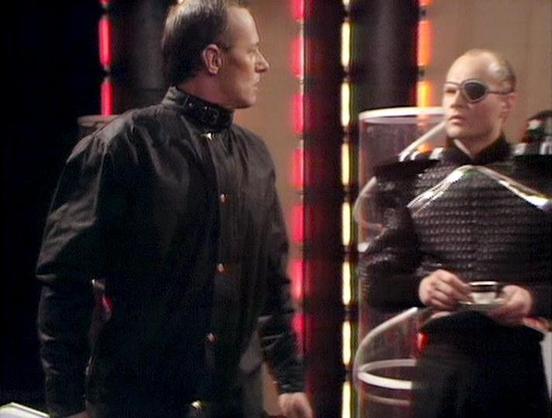 Claude Haiku 4.5 describes the image as: "# Image Description

This image appears to be from a science fiction production, featuring two figures in a futuristic setting. The scene is illuminated by neon lighting in red and yellow tones, creating an atmospheric, stylized environment. The figure on the left wears a black outfit with distinctive collar details, while the figure on the right wears sunglasses and dark textured clothing, holding what appears to be a data pad or similar device.

The s…