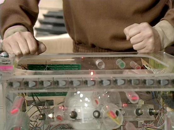 Claude Sonnet 4.5 describes the image as: "This image shows a close-up view of someone interacting with what appears to be a transparent electronic device or experimental apparatus. The person, wearing a brown/tan colored top, is gripping the edges of a clear acrylic or plastic case with both hands. Inside the transparent housing, there's a complex array of electronic components including circuit boards (visible green PCBs), various colored wires, LEDs or indicators (pink/red visible), and what…