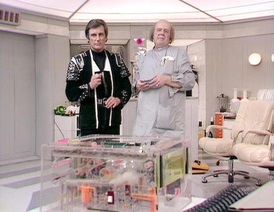 Claude Sonnet 3.7 describes the image as: "This image shows a scene from what appears to be a science fiction TV production, likely from the British sci-fi series "Blake's 7" or a similar program. Two performers are standing in a futuristic-looking white set that resembles a spacecraft or high-tech facility.

The person on the left wears a distinctive black costume with ornate embellishments, while the person on the right is dressed in a light gray or white outfit. Between them is a transparent…