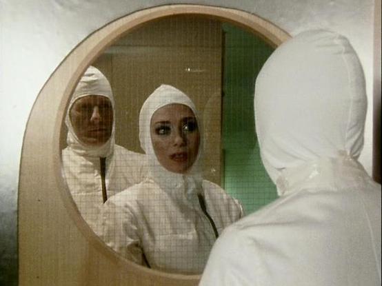 Claude Sonnet 3.7 describes the image as: "The image shows a scene that appears to be from a film or television production, likely from a science fiction or medical drama setting. It features people in white protective suits or medical garments with head coverings, standing in front of a circular mirror or viewing window. The setting has a clinical, sterile appearance with what looks like green tiling visible in the background.

The composition creates an interesting visual effect where we see …