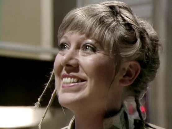 Claude Haiku 4.5 describes the image as: "I can see a woman in what appears to be an indoor setting with neutral-toned walls in the background. She has shoulder-length blonde hair styled in a casual updo with face-framing pieces, and is wearing what looks like a dark-colored top. She's smiling and looking off to the side, appearing engaged and cheerful in the moment. The lighting suggests this is from a television production, with professional studio lighting visible. The setting appears to be …