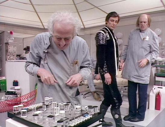 Claude Haiku 4.5 describes the image as: "# Scene Description

This image shows a futuristic laboratory or control room setting with a distinctive geodesic dome ceiling featuring geometric panels. The space features white walls and modern equipment typical of science fiction production design from the 1970s-80s era.

In the foreground, an older man with white hair in a light gray tunic examines what appears to be a detailed miniature model or diorama laid out on a table. The model contains nume…