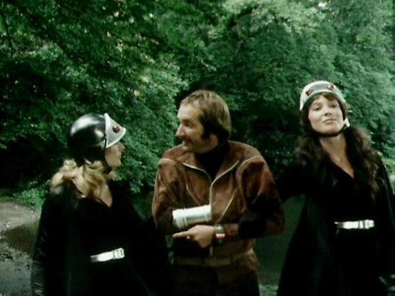 Claude Sonnet 4 describes the image as: "This appears to be a scene from a science fiction production, likely from the late 1970s or early 1980s based on the film quality and costume design. The setting is outdoors in a lush, green wooded area with dense foliage creating a natural backdrop. Three actors are positioned together, wearing what appears to be futuristic or military-style clothing in dark colors. Two of the performers are wearing distinctive headgear or helmets that suggest this is f…