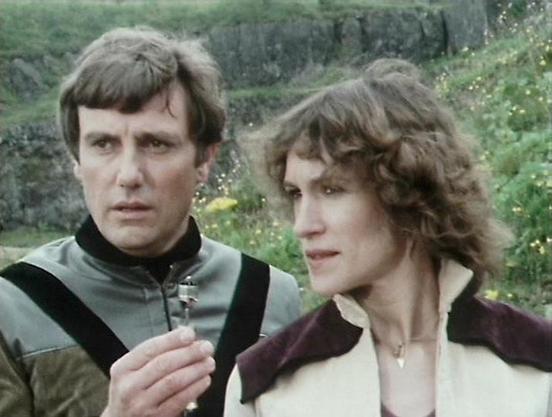 Claude Haiku 4.5 describes the image as: "# Scene Description

This image captures two actors in an outdoor setting with lush greenery and flowering plants visible in the background. The scene appears to be from a science fiction television production, given the distinctive costume design.

On the left, one actor wears a gray and black tunic-style outfit with a cross pendant, displaying a serious, contemplative expression. The actor on the right wears a cream-colored jacket with dark collar det…