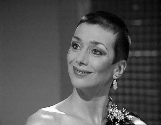 Claude Haiku 4.5 describes the image as: "# Black and White Portrait

This is a classic black and white photograph showing a woman in what appears to be a scene from a mid-20th century film or television production. She is photographed in profile, turning slightly toward the camera with a warm, engaging smile. Her short, dark hair is styled in a typical fashion of that era, and she wears dangling earrings that catch the light.

The woman is dressed elegantly in what appears to be a light-colore…