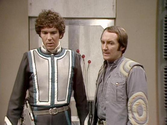Claude Sonnet 3.7 describes the image as: "This image shows a scene from the classic British science fiction television series "Blake's 7." The still features two characters in futuristic costumes standing side by side in what appears to be a spacecraft interior with typical 1970s/early 1980s sci-fi production design.

The figure on the left has curly hair and wears a distinctive white and blue uniform with dark sleeves and a utility belt. The figure on the right has straight hair and is dresse…