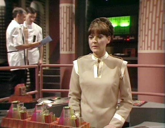 Claude Sonnet 4 describes the image as: "I can see this image shows what appears to be a scene from a science fiction television production, likely from the 1970s or 1980s based on the visual style and set design. The setting appears to be a futuristic command center or spacecraft bridge, with characteristic sci-fi elements like metallic railings, control panels, and monitors visible in the background. Two figures in white uniforms can be seen in the background area.

The scene has the distinct…