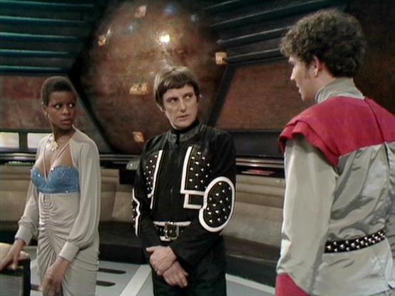 Claude Sonnet 4.5 describes the image as: "This image appears to be from the classic British science fiction television series "Blake's 7," which aired from 1978 to 1981. The scene takes place on what looks like a spaceship set, with a distinctive large spherical object visible in the background that's characteristic of the show's design aesthetic.

Three characters are shown in conversation on the ship's bridge or control room. The character on the left wears an elegant draped costume with a b…