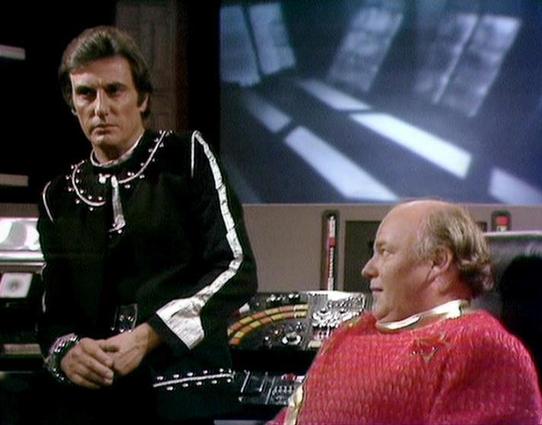 Claude Sonnet 4 describes the image as: "This appears to be a scene from a science fiction television production, likely from the late 1970s or early 1980s based on the visual style and set design. The setting is clearly a futuristic spacecraft or space station, with characteristic sci-fi elements including control panels, monitors, and sleek technological surfaces visible in the background.

The scene shows two men in what appears to be a command center or bridge area. One figure is standing, …