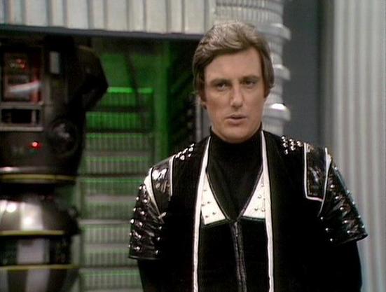Claude Sonnet 4 describes the image as: "This image shows a scene from the British science fiction television series "Blake's 7," featuring Paul Darrow in his iconic role as Kerr Avon. Darrow is wearing Avon's characteristic black leather outfit with metallic studs and white trim details, which became synonymous with the character throughout the series. The setting appears to be aboard the Liberator spaceship, with typical sci-fi set design visible in the background including control panels and…