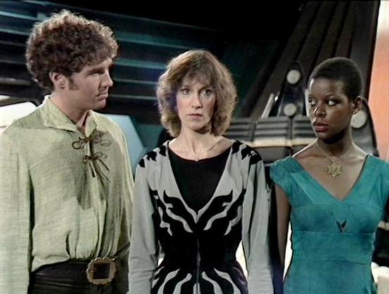 Claude Sonnet 4.5 describes the image as: "This image appears to be from the British science fiction television series "Blake's 7," which aired from 1978 to 1981. The scene shows three actors on what appears to be a spaceship set, characterized by the futuristic interior with angular walls and technical equipment visible in the background.

Steven Pacey stands on the left wearing a beige or cream-colored tunic with distinctive lacing detail at the chest and a dark belt. Jan Chappell is position…