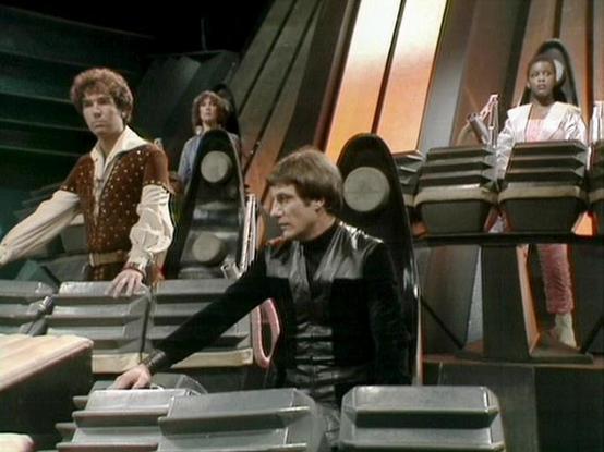 Claude Sonnet 4 describes the image as: "This image shows a scene from what appears to be a science fiction television production, likely from the late 1970s or early 1980s based on the production values and costume design. The setting is a futuristic spacecraft or space station interior, featuring characteristic angular metallic walls, multi-level platforms connected by stairs, and ambient lighting that creates an otherworldly atmosphere.

The scene captures four actors positioned at different…