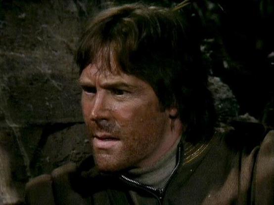 Claude Sonnet 3.7 describes the image as: "This image appears to be a still from a film or television production, showing a person with brown hair in what looks like a rugged setting. The individual is wearing what appears to be dark clothing with some kind of zipper detail visible. The background suggests a stone or rock wall environment, creating a somewhat gritty, possibly historical or adventure-themed atmosphere.

The lighting is dramatic and somewhat dim, giving the scene a serious tone. …