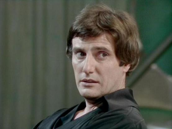 Claude Sonnet 4 describes the image as: "This image shows a dramatic scene featuring a man with dark, wavy hair wearing what appears to be a black leather or similar dark garment. The setting has a futuristic or sci-fi aesthetic, with green-tinted lighting and metallic structural elements visible in the background. The lighting creates a moody, atmospheric effect typical of science fiction television productions from the late 1970s or early 1980s. The man's expression appears intense or concern…