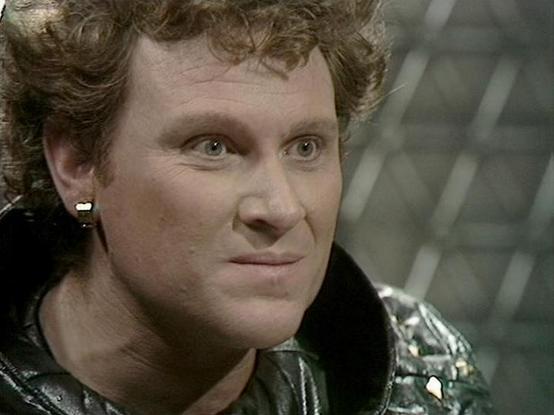 Claude Sonnet 3.7 describes the image as: "This image appears to be from a science fiction TV production, likely from the 1980s based on the visual style and quality. The person in the image has curly dark hair and is wearing what looks like a distinctive costume with some metallic or reflective elements on the shoulders. They have a subtle expression that suggests a moment of contemplation or mild reaction to something happening off-screen.

The background is somewhat blurred but appears to be…