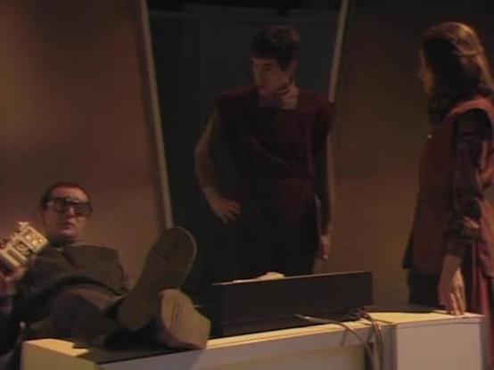 Claude Sonnet 3.7 describes the image as: "The image shows a scene from what appears to be a TV show or film with a professional office setting. There are three people in the frame. On the left, a person in glasses wearing a suit is seated with their feet propped up on what looks like a desk, holding what might be papers or a magazine. Standing in the middle and on the right are two other individuals in dark reddish-brown attire, appearing to be in conversation or a meeting situation.

The ligh…