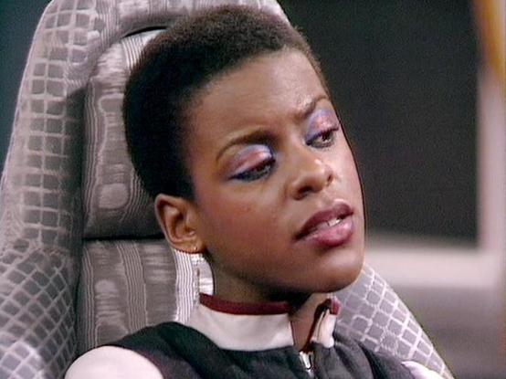 Claude Sonnet 3.7 describes the image as: "The image shows a person with short dark hair wearing colorful eye makeup and lipstick. They appear to be seated in a chair with a patterned gray upholstery. The person is wearing what looks like a uniform or costume with a black body and white collar detail.

The setting appears to be from a television or film production, with the individual positioned in what seems to be an interior set. The lighting and film quality suggest this is from a production…