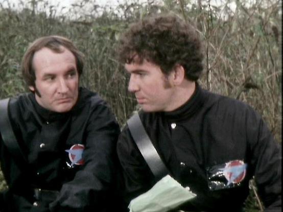 Claude Sonnet 3.7 describes the image as: "The image shows two individuals in a scene from what appears to be a science fiction television production. They are wearing identical black uniforms with distinctive circular emblems featuring a red and blue design on the chest. One person has straight hair while the other has curly hair. They appear to be having a conversation in an outdoor setting with dried vegetation or reeds in the background, suggesting a wilderness or remote location.

The styl…