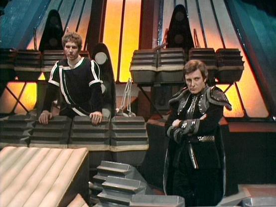 Claude Sonnet 4.5 describes the image as: "This image shows a scene from the British science fiction television series "Blake's 7" (1978-1981). The setting appears to be aboard the Liberator spaceship, recognizable by its distinctive multi-colored curved panels and geometric architecture in the background, featuring orange, yellow, and blue hues.

Two characters are positioned on different levels of the set's angular stairway design. On the left, Steven Pacey portrays Del Tarrant, wearing a bla…