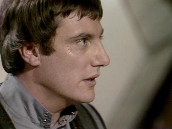 Claude Haiku 4.5 describes the image as: "# Image Description

This is a profile shot of a man shown in what appears to be a scene from a television production, likely from the 1970s based on the image quality and styling. The man is captured in a side profile view, facing toward the right of the frame. He has dark brown hair styled in a typical 1970s fashion with slight waves and length at the sides. His facial features are sharp and defined, with a distinctive nose and angular jawline.

He's …