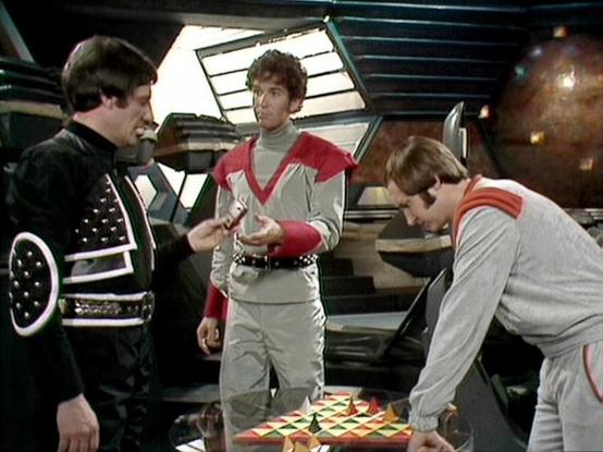 Claude Haiku 4.5 describes the image as: "# Scene from Blake's 7

This image shows a scene from the science fiction television series "Blake's 7," set aboard a spacecraft interior with futuristic design elements. The setting features distinctive 1970s sci-fi aesthetics with curved panels, illuminated skylights, and control room furnishings typical of the show's production design.

Three characters are gathered around what appears to be a tactical or gaming table with a geometric pattern display…