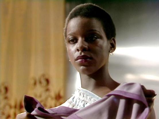 Claude Sonnet 3.7 describes the image as: "This image shows a scene that appears to be from a period drama or film. The person in the foreground is wearing an elegant outfit with a white top that has detailed embroidery or lace at the collar, paired with what looks like a lavender or light purple fabric draped over the shoulders. 

The background suggests an interior setting with warm-toned walls, possibly in a historical building or set designed to evoke a certain era.

The composition of the …