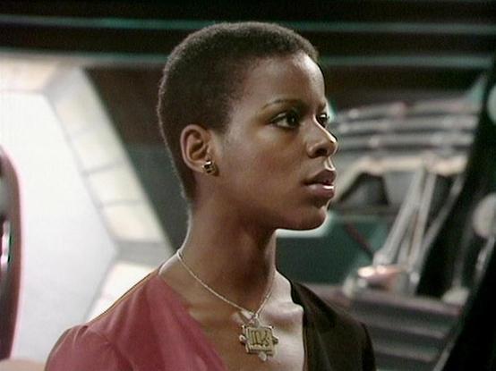 Claude Haiku 4.5 describes the image as: "# Image Description

This image shows a scene set in a futuristic spacecraft interior, characterized by metallic walls and clinical lighting typical of science fiction settings. The person in the frame is positioned in profile, wearing a burgundy/maroon colored top paired with a distinctive metallic pendant necklace featuring geometric patterns. The styling, including the short hair and wardrobe choices, reflects the aesthetic of 1990s science fiction t…