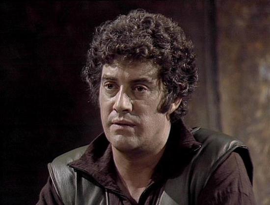 Claude Sonnet 4 describes the image as: "This image shows a man with dark, curly hair wearing what appears to be a brown or dark-colored leather jacket or tunic. The setting has a dimly lit, atmospheric quality with shadowy backgrounds that suggest this is from a television production or film, likely from the 1970s or 1980s based on the image quality and styling. The lighting creates a dramatic, moody atmosphere typical of science fiction productions from that era. The costume and production va…