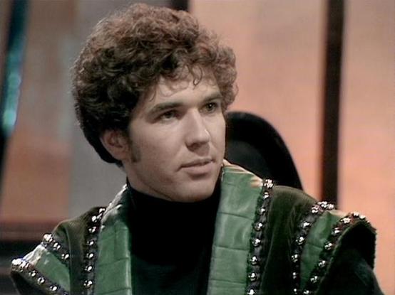 Claude Sonnet 4.5 describes the image as: "This image shows a scene from what appears to be a British science fiction television production, likely from the late 1970s or early 1980s based on the video quality and styling. The actor Steven Pacey is wearing a distinctive costume consisting of a dark green quilted leather or vinyl vest with metallic studded trim along the edges, paired with a black turtleneck underneath. His hair is styled in a characteristic curly, voluminous fashion typical of …