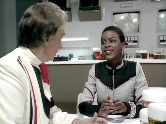 Claude Sonnet 3.7 describes the image as: "This image appears to be from a classic science fiction TV show, likely from the 1980s based on the production style and set design. The scene takes place in what looks like a futuristic control room or spaceship interior with technical equipment visible in the background.

Two people are engaged in conversation. On the left is a person in a distinctive white outfit with red and black accents. On the right is someone wearing a sporty black and white ja…