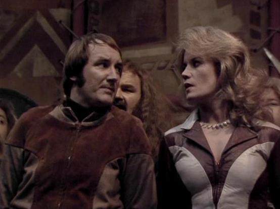 Claude Sonnet 4.5 describes the image as: "This image appears to be from the British science fiction television series "Blake's 7," which aired from 1978-1981. The scene shows three characters in what looks like a spaceship or futuristic interior setting, with angular metallic panels visible in the background.

On the left is a male character wearing a distinctive brown and black costume with a high collar, sporting dark hair with a somewhat tousled style. To the right is a female character wit…