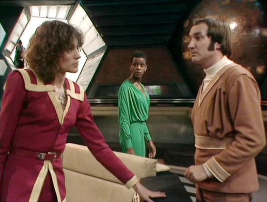 Claude Sonnet 4 describes the image as: "This appears to be a scene from the British science fiction television series "Blake's 7" from the late 1970s or early 1980s. The setting is clearly a futuristic spacecraft interior, with characteristic hexagonal viewports and sleek control panels visible in the background showing stars in space.

The scene shows three characters in a tense conversation aboard what appears to be the Liberator spacecraft. On the left is a woman with curly dark hair wearin…