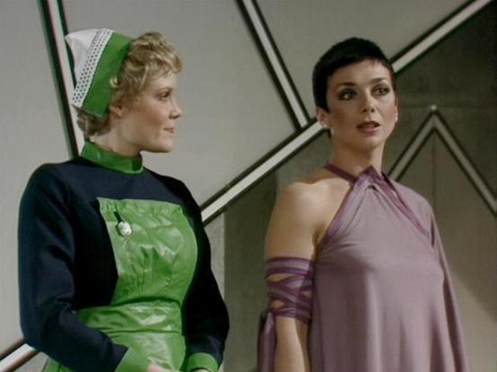 Claude Haiku 4.5 describes the image as: "# Scene Description

This image captures two women in what appears to be a futuristic interior setting, characterized by sleek geometric wall panels and a minimalist design aesthetic typical of 1970s science fiction production design.

On the left stands a woman wearing a striking green and black ensemble consisting of a black long-sleeved top paired with a bright green quilted tunic or dress. She accessorizes with a coordinating green headband featurin…