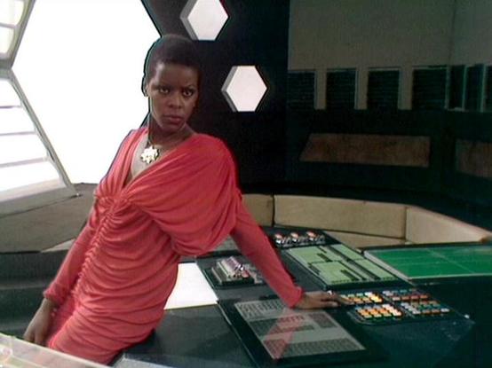 Claude Haiku 4.5 describes the image as: "I can see a woman in a red draped top standing at what appears to be a control panel or desk in a sci-fi setting. The background features modern, minimalist design elements including hexagonal lighting panels on the wall and a curved architectural style typical of futuristic set design. She's positioned at what looks like a technical console with various controls and displays. The lighting is bright and clinical, contributing to the high-tech atmosphere…