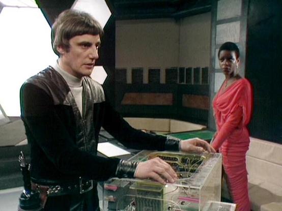 Claude Sonnet 4 describes the image as: "This appears to be a scene from the British science fiction television series "Blake's 7," likely from the 1980s based on the production style and set design. The image shows two characters in what appears to be a futuristic spacecraft or space station setting. One character is wearing a distinctive black leather outfit with a high collar, positioned at what looks like a control console or computer terminal with transparent components. The other characte…