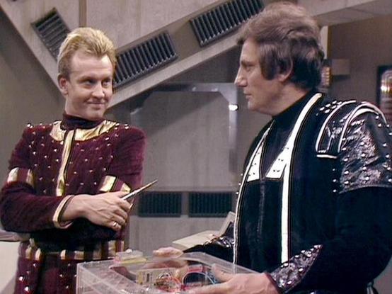 Claude Haiku 4.5 describes the image as: "# Scene Description

This image captures a scene from what appears to be a science fiction television production, set in a futuristic interior with metallic architectural elements visible in the background. The setting features modernist design with horizontal vents and sleek surfaces typical of 1970s-80s sci-fi aesthetics.

Two male actors are engaged in conversation. The actor on the left wears an ornate burgundy tunic with gold trim and embellishment…