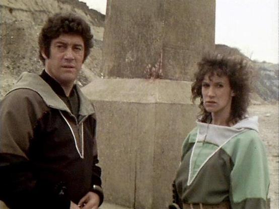 Claude Sonnet 4.5 describes the image as: "This image appears to be from a science fiction television production, likely from the late 1970s or early 1980s based on the visual quality and styling. The scene is set in what looks like a quarry or industrial location with concrete structures and rough terrain in the background.

Two figures stand in the frame wearing distinctive costumes that suggest a futuristic or space-faring setting. The person on the left wears a brown and black outfit with w…