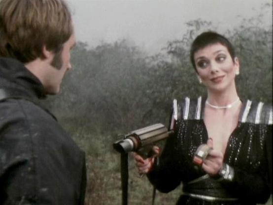 Claude Sonnet 4 describes the image as: "In this atmospheric scene from the British sci-fi series Blake's 7, two characters are engaged in conversation in what appears to be a misty, wooded outdoor setting. The male character on the left wears a dark leather jacket and has his back partially to the camera. The female character faces him, dressed in a striking black outfit with metallic studs or decorations and white straps, giving her a distinctive, futuristic appearance. She has dark hair styl…